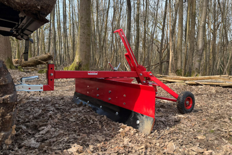 Red log splitter in a leaf-covered forest clearing, with a wedge and hydraulic arms and bare trees in the background.
