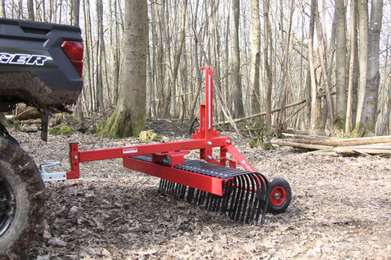 Red forestry rake attachment on a trailer in a leaf-covered forest; bare trees in the background.