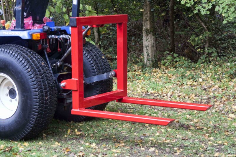 Red metal frame of an agricultural implement attached to a tractor, outdoors among trees and fallen leaves.