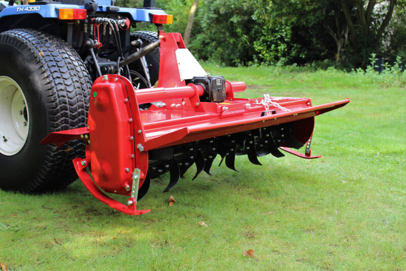 Red farm implement attached to a blue tractor, with green field in the background.