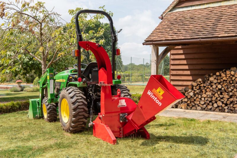 Green John Deere tractor with red wood chipper attachment beside a stacked log pile near a wooden shed in a yard
