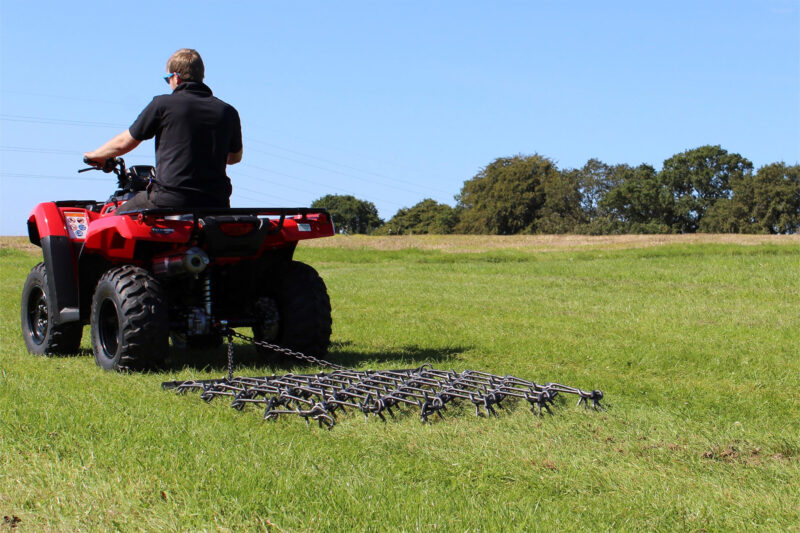 Person on a red ATV riding across a grassy field under a blue sky, wearing a black shirt and sunglasses (side view).