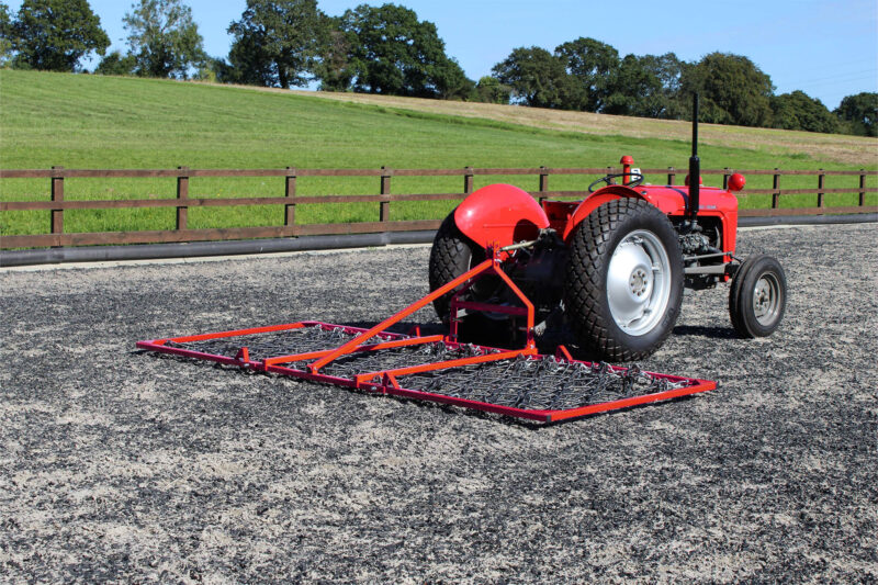 Red tractor with a large cultivator attachment on a gravel surface, green fields and trees in the background behind a wooden fence.