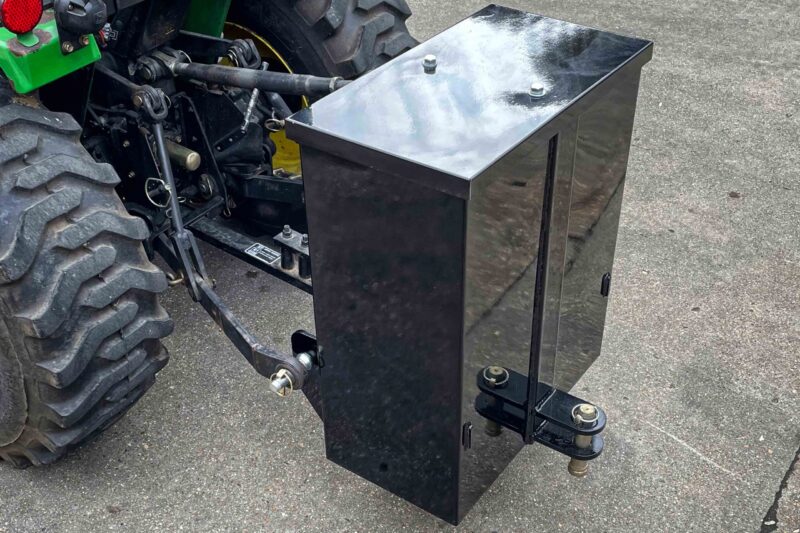 Black metal toolbox mounted on a tractor hitch with bolts and hydraulic links nearby.