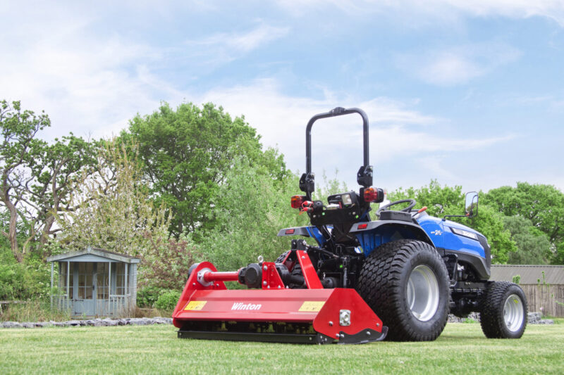 Blue tractor with a red mower attachment on a green lawn, trees in background under a clear sky.