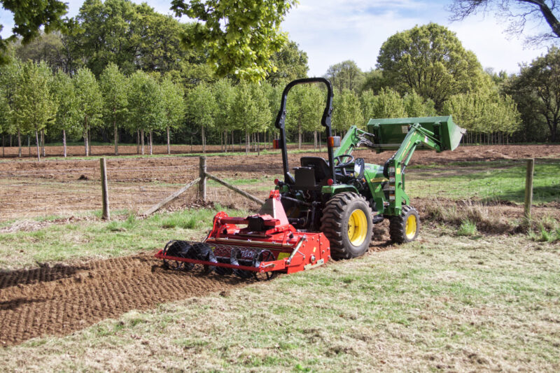Green tractor with a red tiller attachment on freshly plowed brown soil, an orchard of young trees in the background under a blue sky.