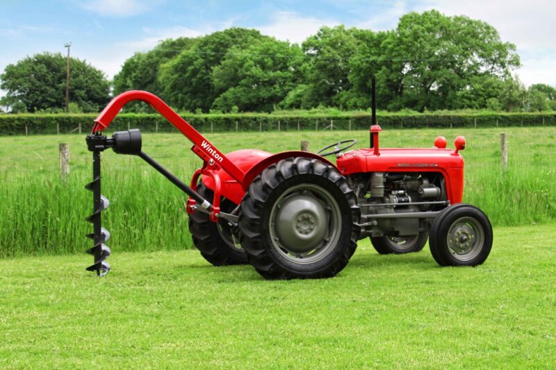 Red farm tractor with a large black auger attachment on a sunny, grassy field; trees and a fence in the background.