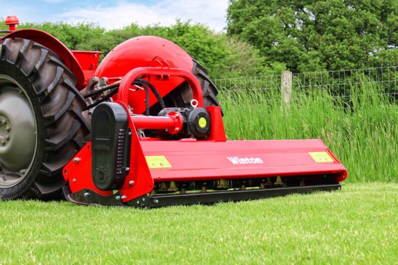 Red agricultural mower attachment on a tractor in a grassy field with a wire fence and green trees in the background, close-up view of machinery components.