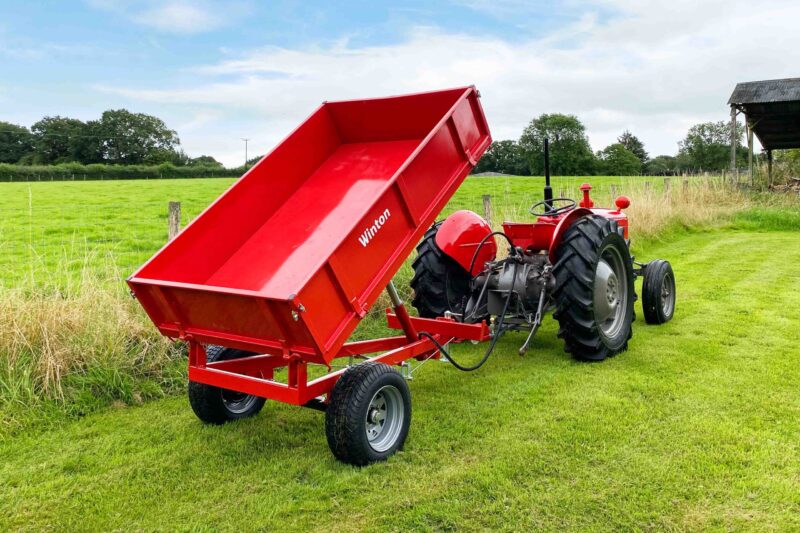 Red farm tractor with a tipped red dump trailer in a green field under a blue sky.
