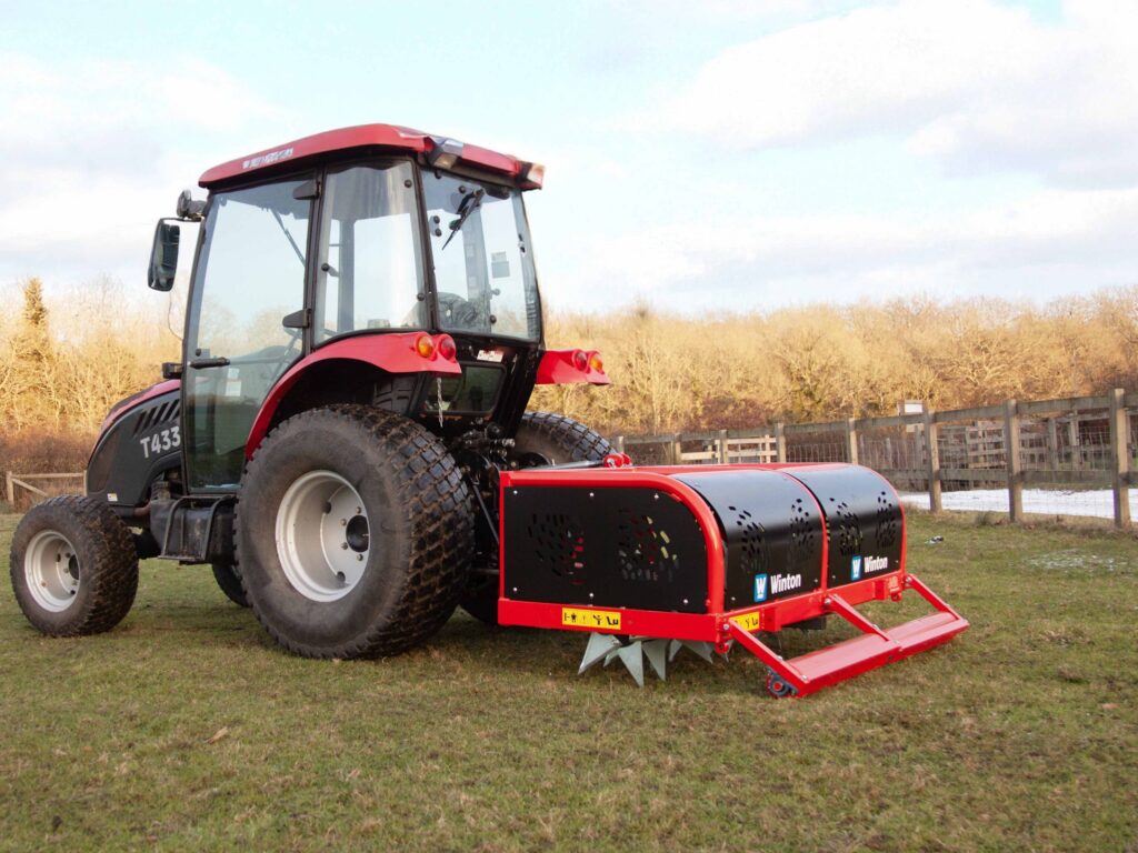 Red tractor with large tires on a grassy field, connected to a red farming implement in a rural area with a wooden fence in the background.