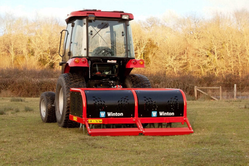 Red tractor with a large front-mounted mower attachment in a grassy field, bare trees in the background