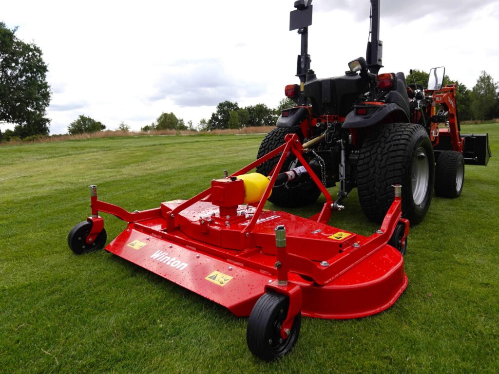 Red heavy-duty mower attachment on a tractor in a grassy field, with large tires and metal hitch visible.