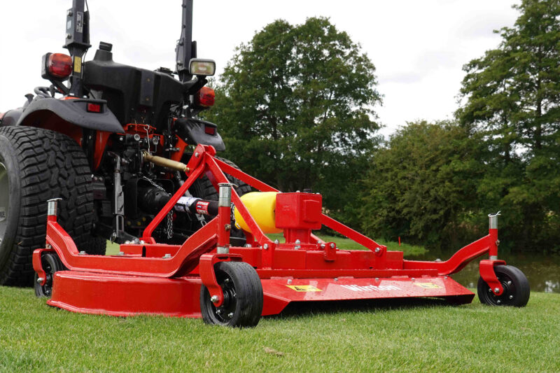 Red agricultural mower attached to a trailer on a grassy field with trees behind.