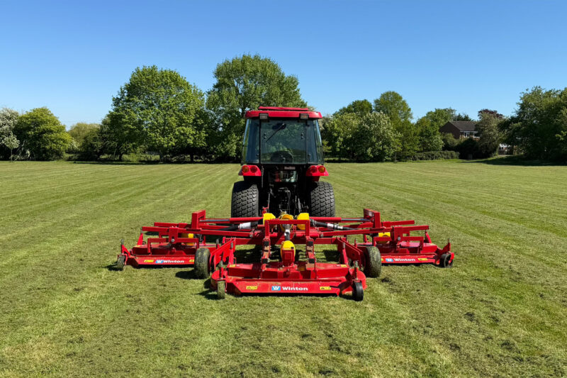 Red tractor with attached red farming equipment on a green field under a clear blue sky, trees and a house in the background.