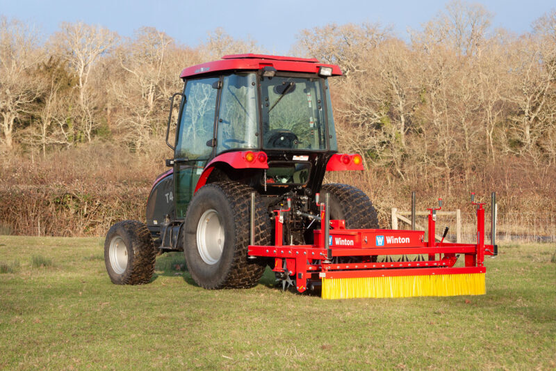 Red tractor with a yellow brush-bar attachment in a grassy field, leafless trees in the background tailing a clear blue sky.