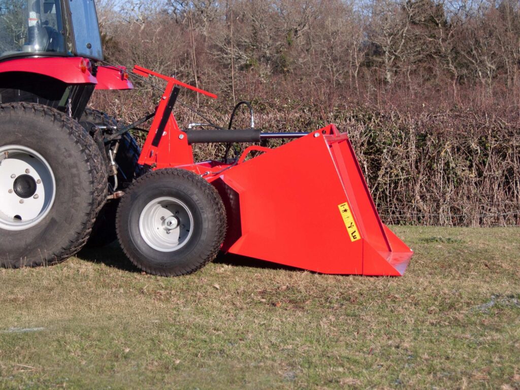 Red tractor with a large front loader attachment on a grassy field, with a leafless hedge in the background.