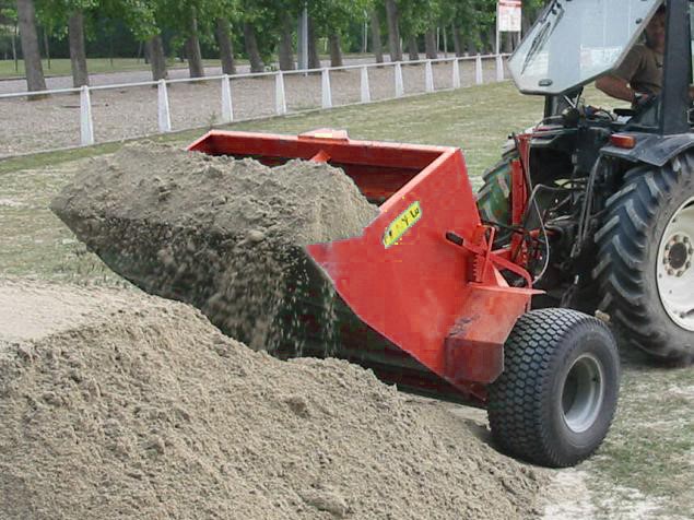 Red dump trailer attached to a tractor dumping sand into a large pile on a field with trees in the background.