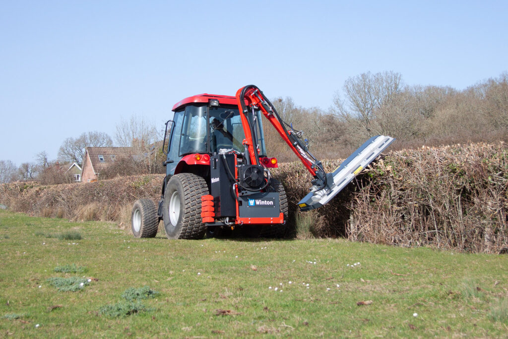 Red heavy-duty tractor-like machine with a hydraulic arm and loading ramp in a grassy, hedge-lined field under a clear blue sky.