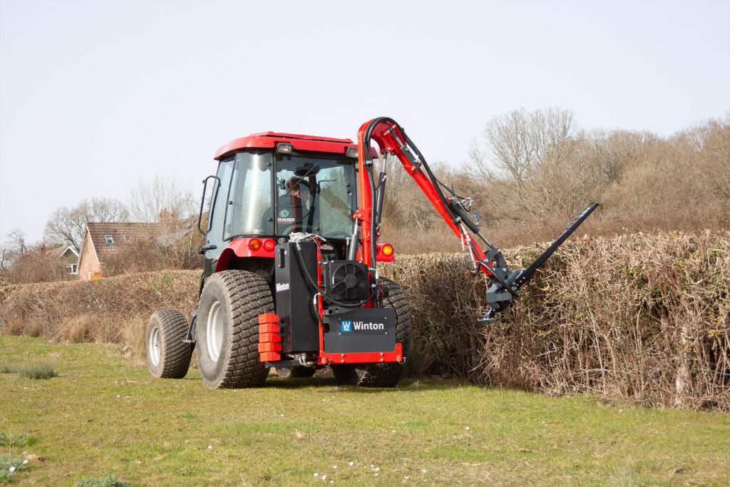 Red agricultural tractor with a hydraulic arm attachment in a grassy field, bare trees and a hedge in the background