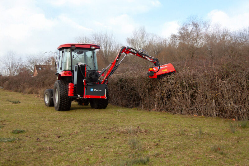 Red tractor with a mounted brush cutter in a grassy field beside a bare-branched hedge in the background.