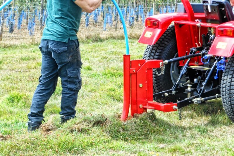 Person in teal shirt and dark cargo pants stands beside a red farming machine in a grassy field. A blue hose is connected to the equipment nearby in an orchard.
