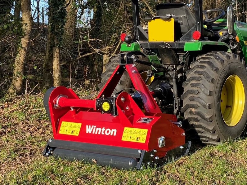 Tractor with a red Winton ground-care attachment (cultivator/tiller) connected for field work in a grassy, wooded area