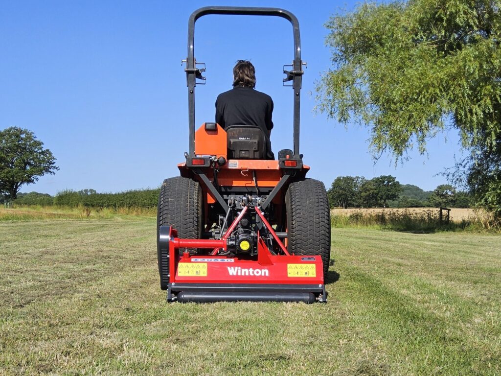 Back view of a person in a black shirt seated on a red Winton agricultural machine in an open field under a blue sky with trees in the background.