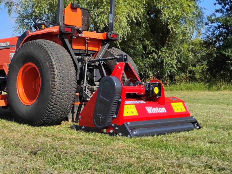 Red Kubota tractor with a large rear attachment on a sunlit grassy field, trees in the background.