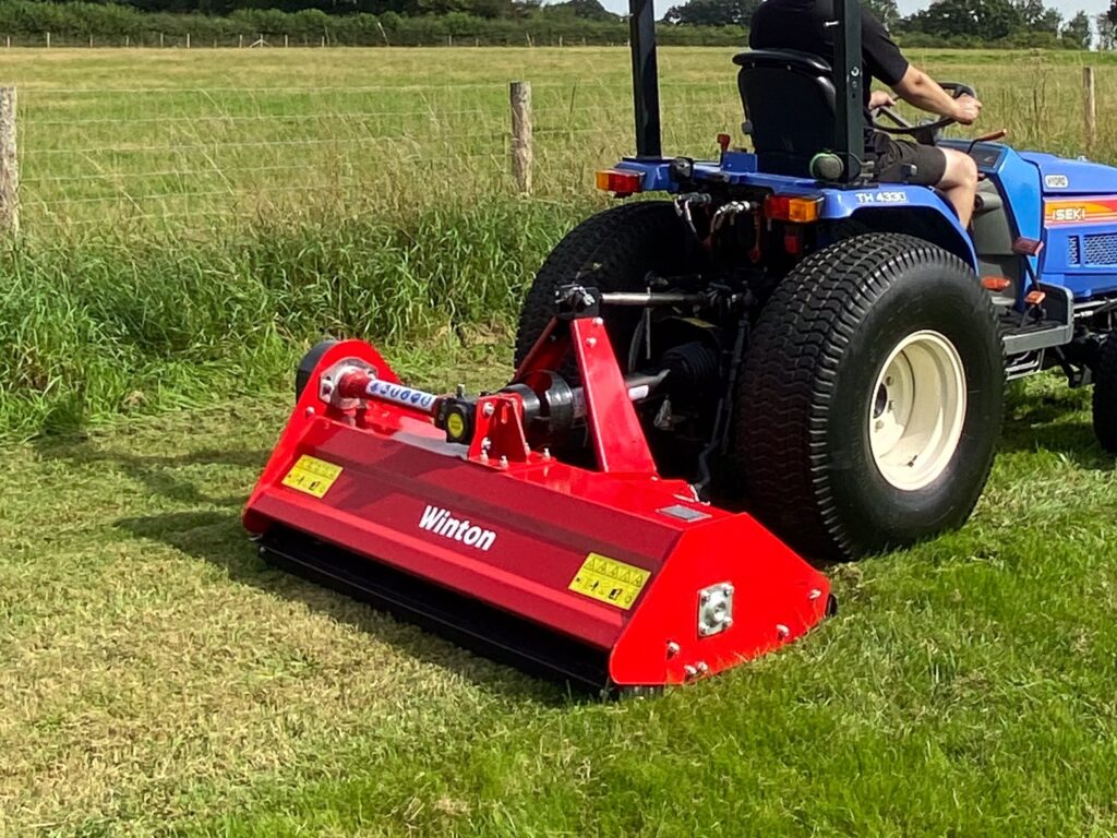 Blue tractor with a large red mower attachment cutting grass in a rural field, driven by a person in a black shirt.