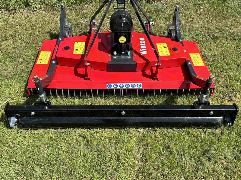 Red lawn mower attachment with rotating tines and warning labels, resting on grass.