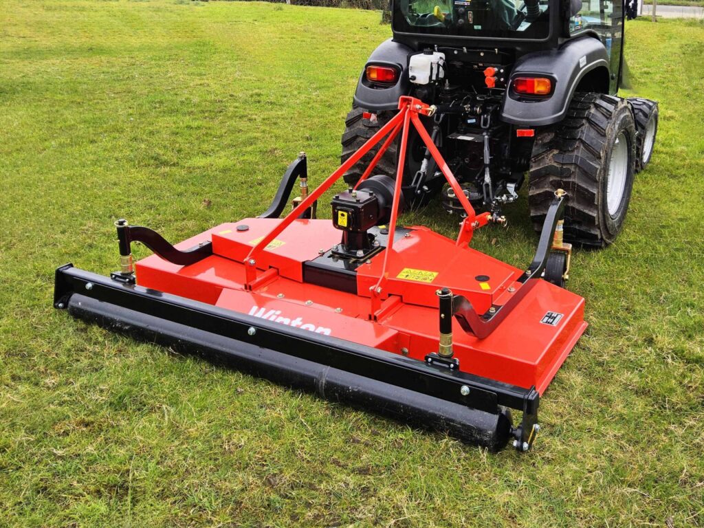 Rear view of a red mower attachment hitched to a tractor on a grassy field.