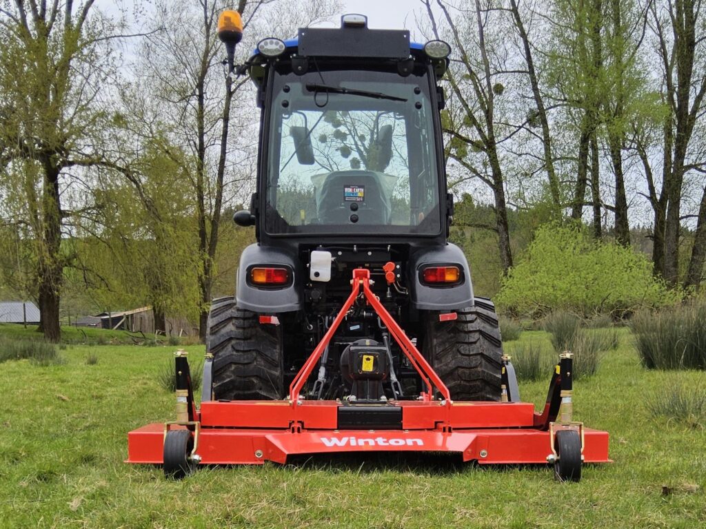 Front view of a large tractor with a red implement attached, orange warning light on top, in a grassy field with trees in the background (Winton branding visible).