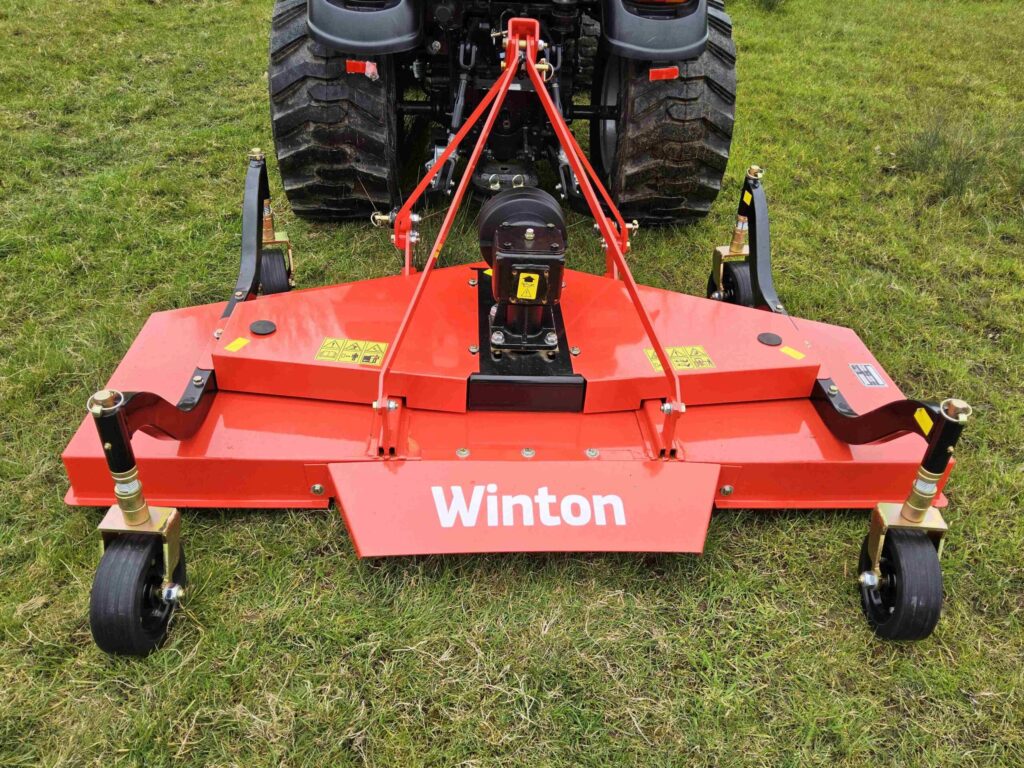 Rear view of a red farming implement attached to a tractor, parked on grass; yellow warning stickers are visible on the equipment.