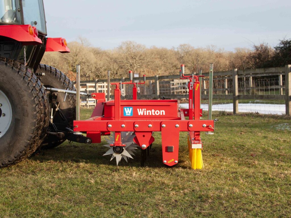 Red farming implement attached to a tractor in a grassy field, with a wooden fence and leafless trees in the background.