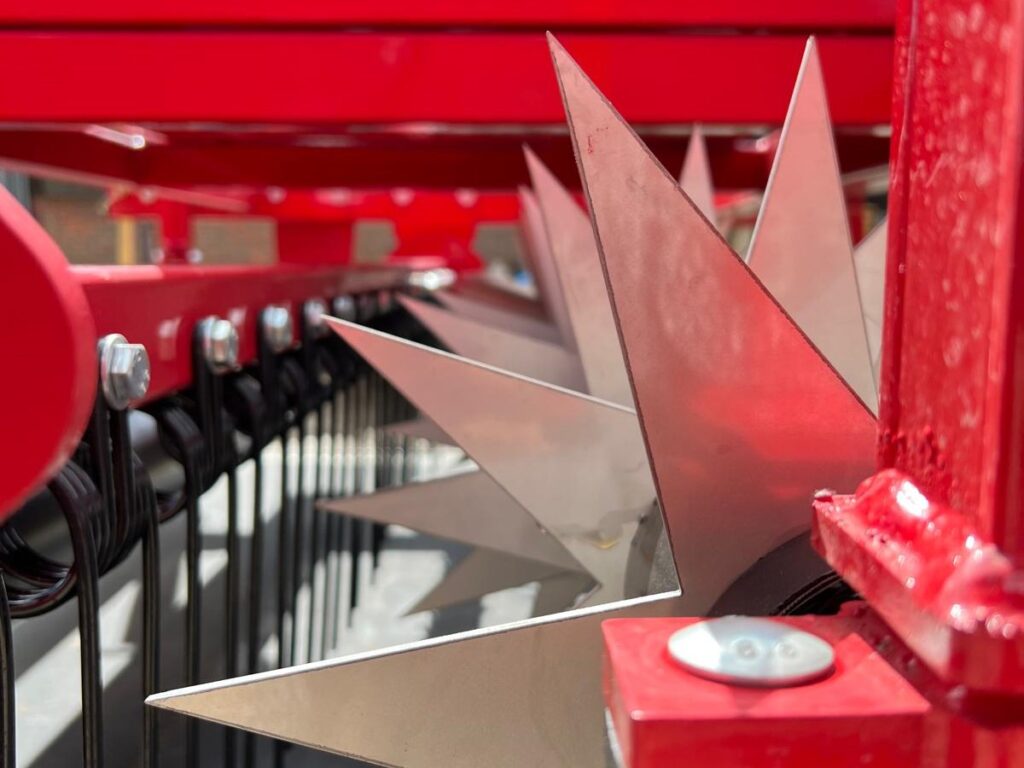 Close-up of red farming machinery with sharp stainless steel blades extending outward, likely part of a mower or harvester.