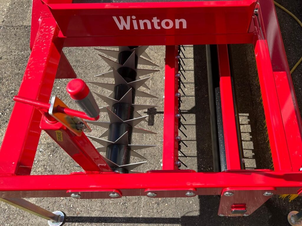 Red Winton farming implement with a rotating auger and sharp, triangular blade tines on a red frame on pavement, close-up view.
