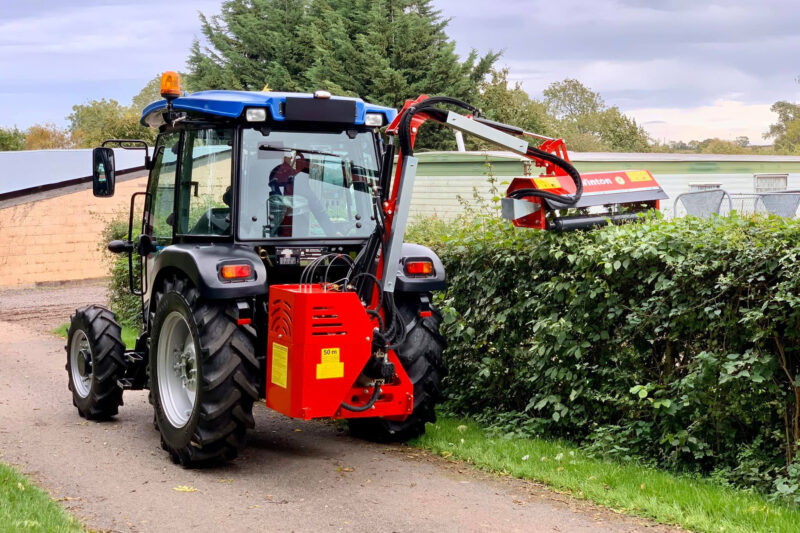 Blue tractor with orange beacon and a red hedge-trimming attachment along a hedged sidewalk area.