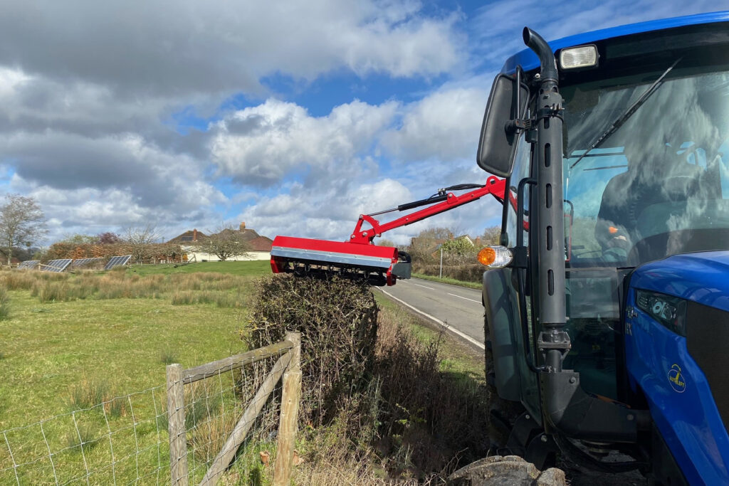 Blue tractor cab with a red hydraulic implement extending toward a fence in a rural field under a cloudy sky and scattered solar panels in the distance.