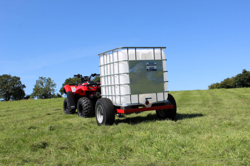 Red off-road utility vehicle towing a large white water tank on a metal-framed trailer in a sunny grassy field under a blue sky.
