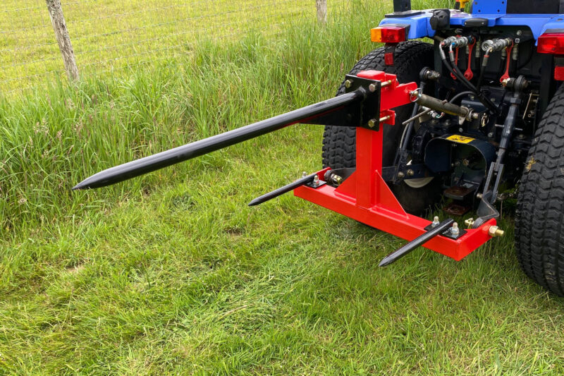 Red tractor attachment with long black spear-like tines extending into tall grass in a field.