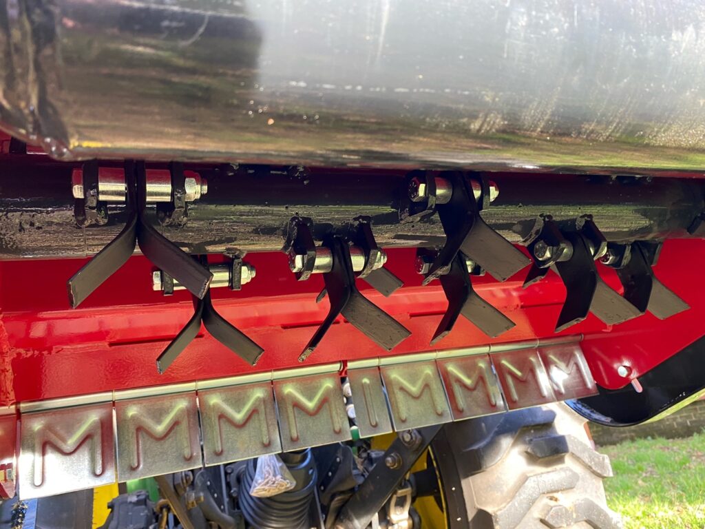 Close-up of the underside of a red agricultural machine showing metal blades, clamps, and hydraulic hoses.