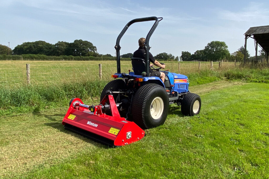 Blue tractor with a large red mower attached, being driven across a grassy field; rural farmland in background.