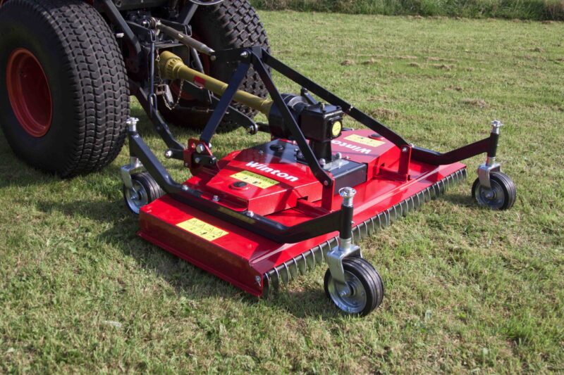 Tow-behind red mower deck attached to a tractor with large off-road tires on a grassy field, ready for mowing down.