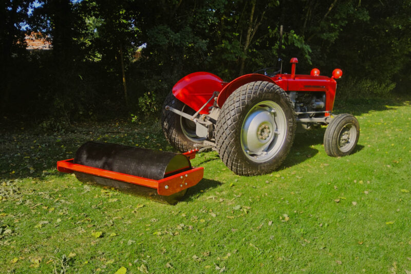 Red tractor with a large black roller attachment on a grassy lawn, trees in the background.