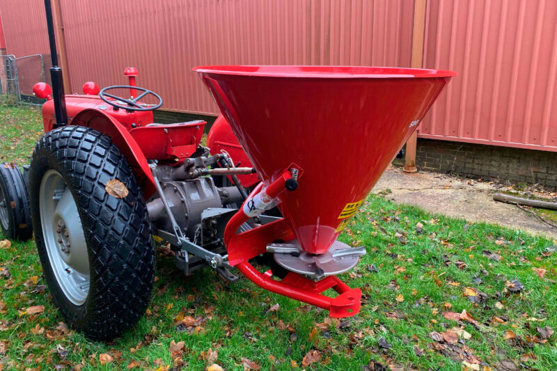 Red tractor with a large red conical hopper attached, parked on grass beside a pinkish-red corrugated wall.