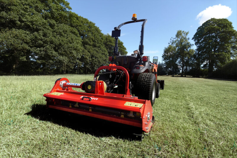 Red agricultural mower attached to a tractor in a sunlit grassy field with trees in the background, under a blue sky