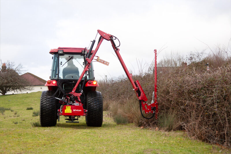 Red tractor with large tires and attached red farming implement in a grassy field, leafless bushes and houses in the background.