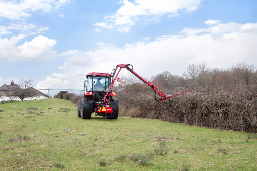 Red tractor with a long hydraulic arm cutting hedges in a field under a blue sky with scattered clouds; houses visible in the distance on the left.