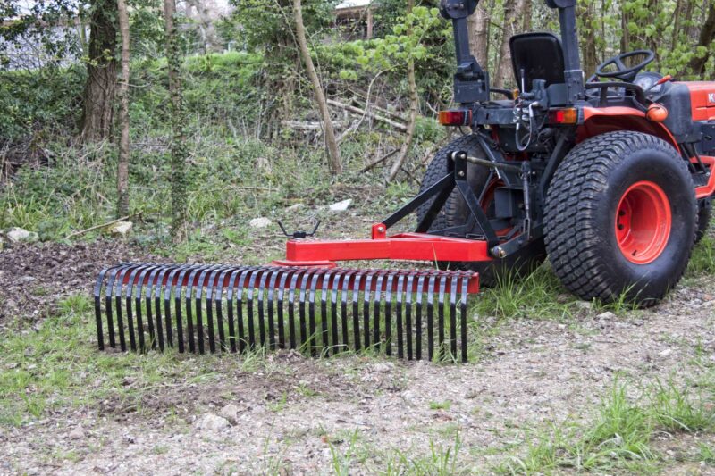 Red tractor with a large front-mounted rake attachment in a wooded, overgrown yard area.