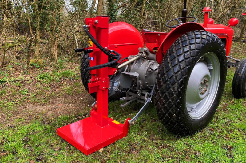 Red agricultural implement attached to a tractor in a grassy, wooded area beside a dirt path.